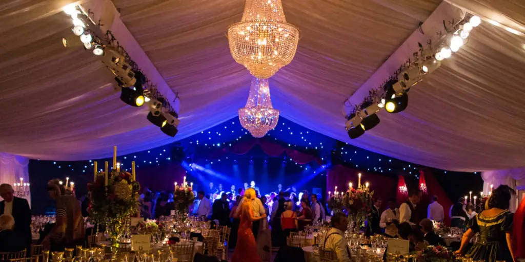 Dine | Exclusive Weddings & Events in Yorkshire | A lively Yorkshire wedding reception in a tent adorned with chandeliers. Guests dance on the floor under blue and purple lights. Elegant tables are set with candles and floral arrangements, creating a festive atmosphere perfect for this luxury event.