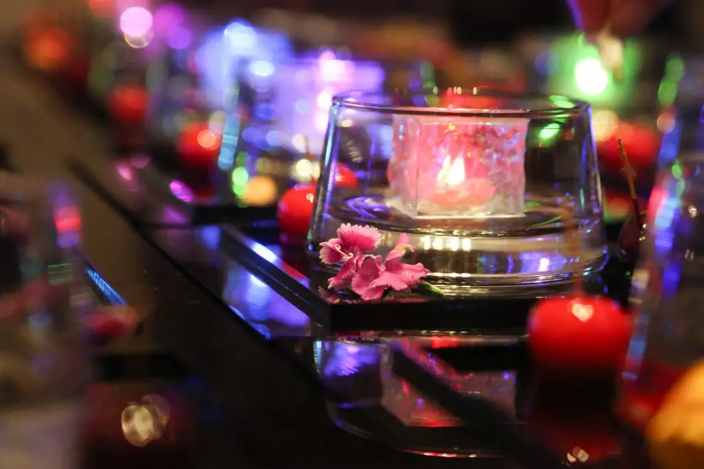 Dine | Exclusive Weddings & Events in Yorkshire | A close-up of a glowing candle in a glass holder on a reflective surface, surrounded by small red balls and a pink flower. Colorful lights create a vibrant, festive atmosphere reminiscent of an elegant Yorkshire wedding.