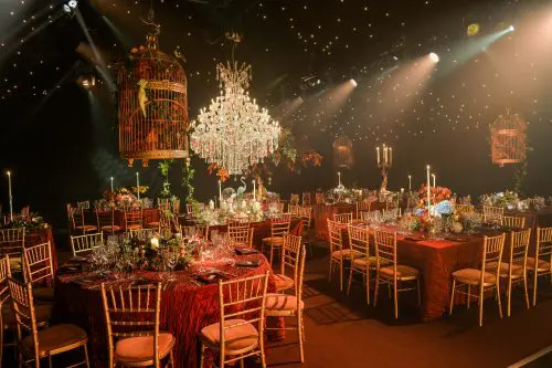 Dine | Exclusive Weddings & Events in Yorkshire | An elegant banquet hall perfect for unforgettable events, featuring tables covered in rich red tablecloths and adorned with floral centerpieces and gold chairs. The sparkling chandelier and hanging birdcages complement the luxurious decor, ideal for a Yorkshire party.