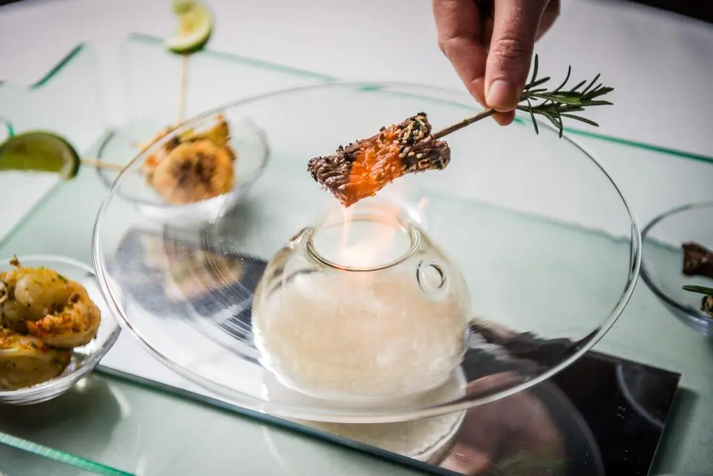 Dine | Exclusive Weddings & Events in Yorkshire | A hand gracefully holds a rosemary sprig skewer with meat over a glass server containing a flame, evoking a sense of luxury dining. The surrounding dishes feature roasted potatoes and garnished seafood, while lime wedges add a vibrant touch to this exquisite food display perfect for events.